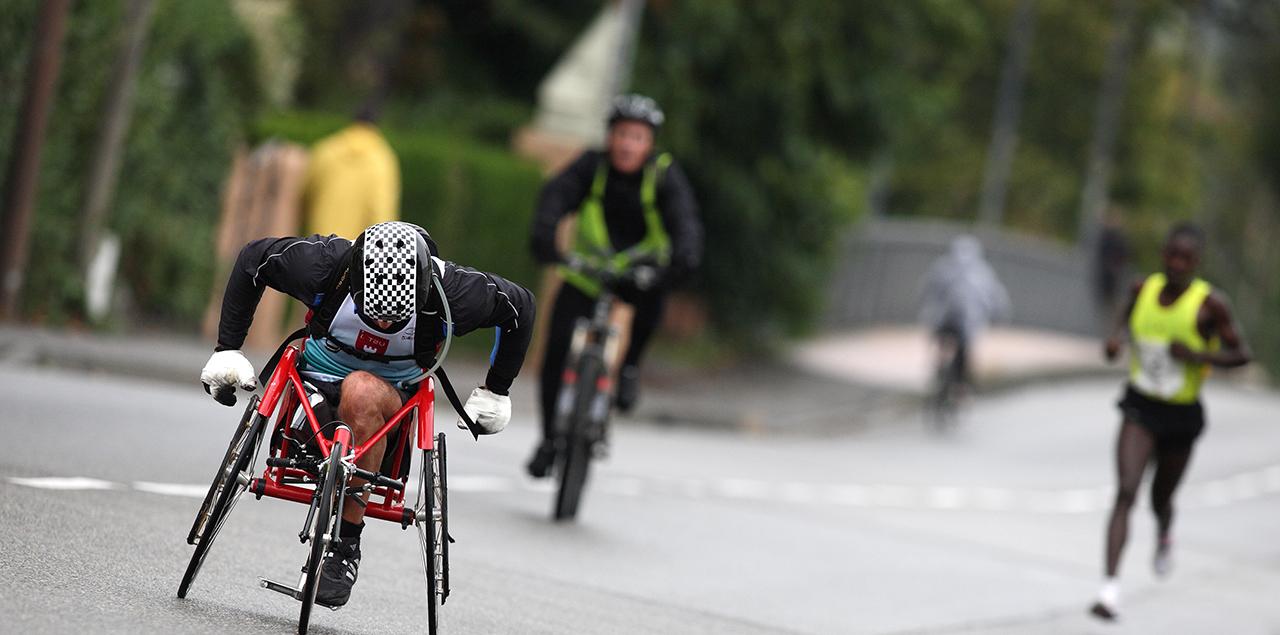 A wheelchair cyclist, a person running and a person riding a bicycle together on an outdoor road