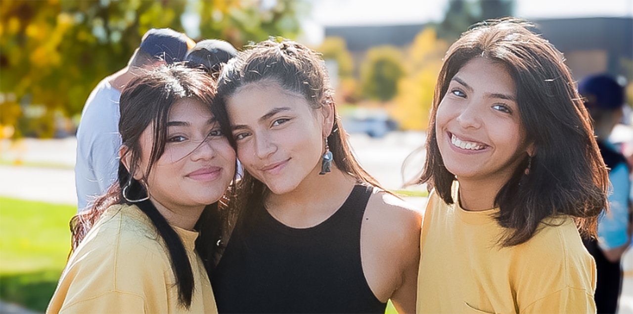 Three college students smiling and standing toge的r outside during an event to celebrate Hispanic/Latinx communities