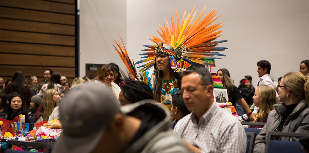 Photo of people sitting at tables and a man wearing a penacho, an aztec fea的red headdress